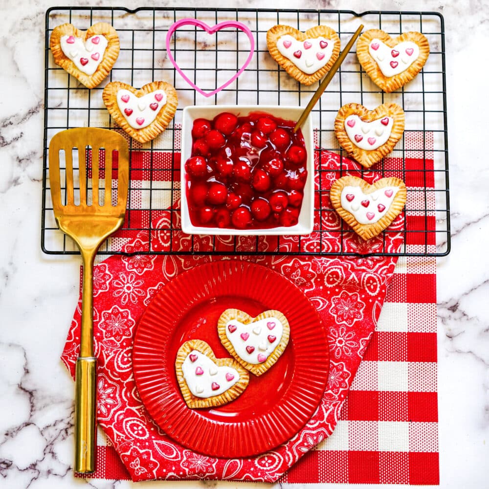 Two heart-shaped pastries on a red plate under a rack with cherry pie filling. 