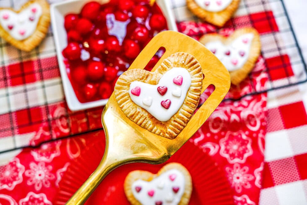 A gold spatula holding a heart-shaped pastry with Valentine's Day frosting and sprinkles. 