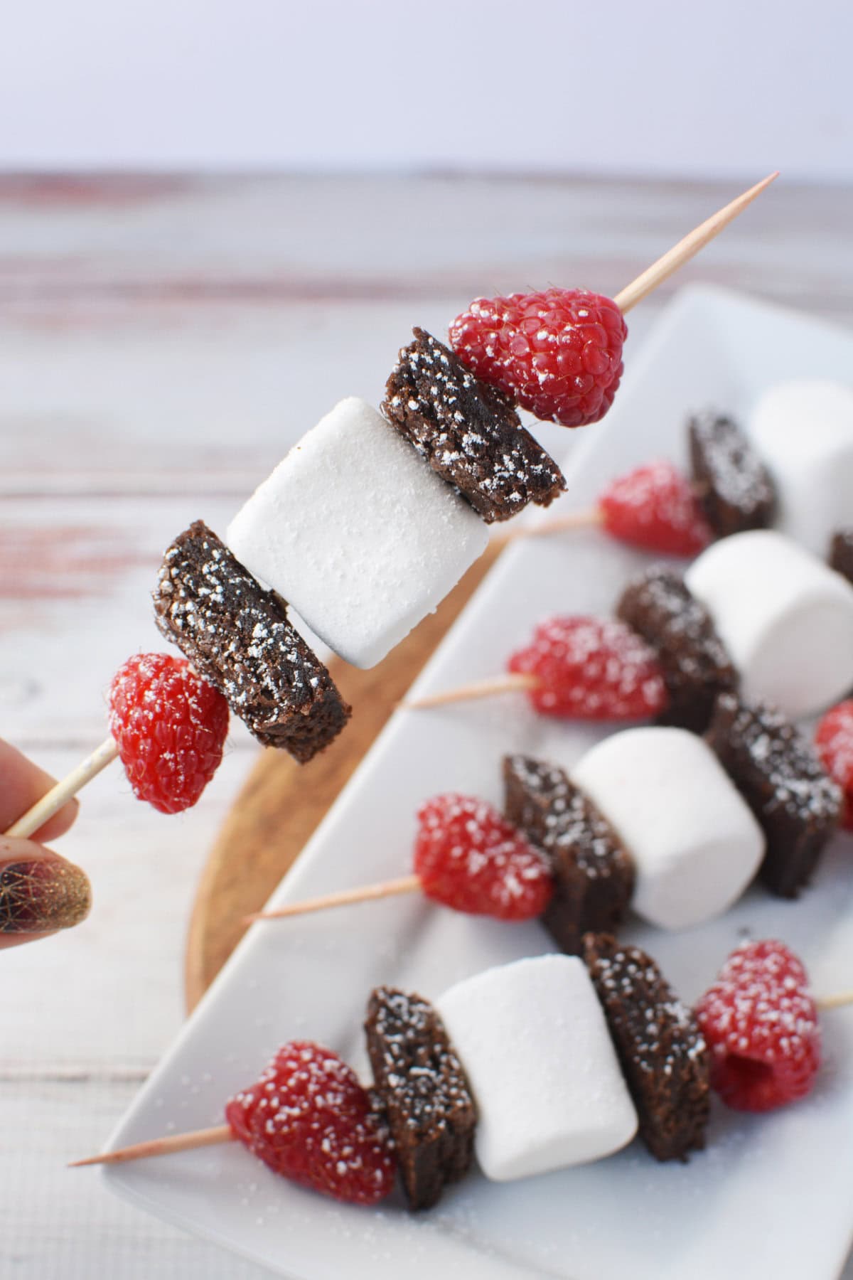 Holding a brownie skewer with brownie, raspberry, and marshmallow.