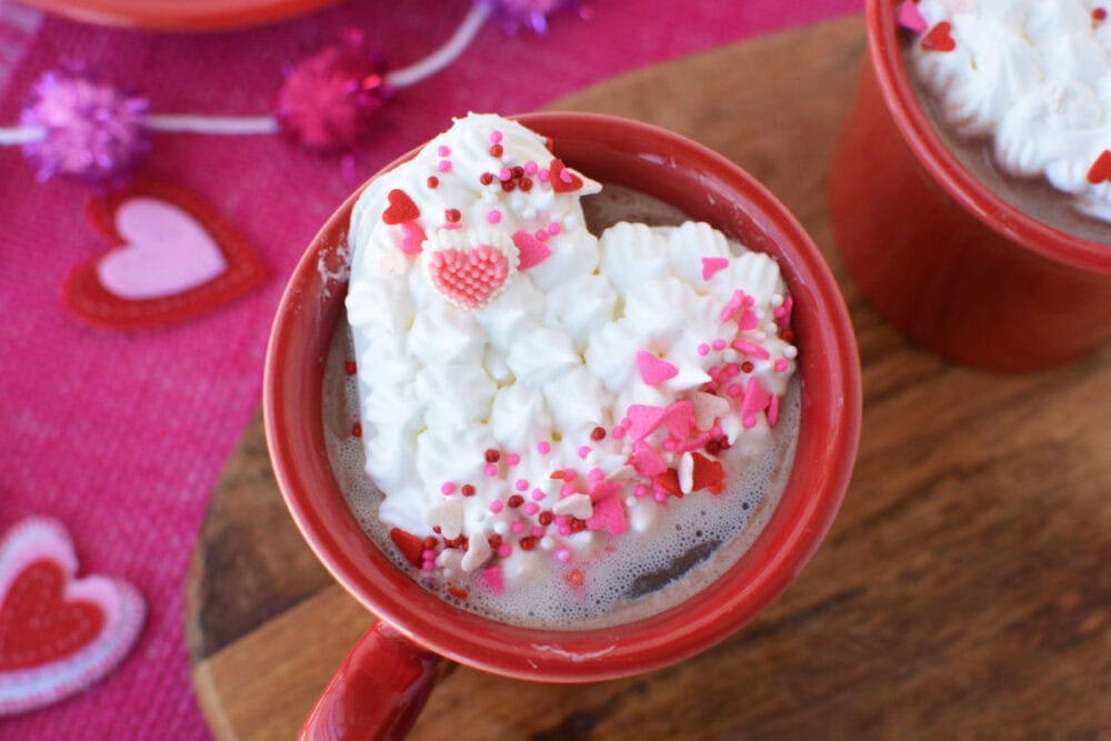 Mug of hot chocolate topped with whipped cream in the shape of a heart with Valentine's Day sprinkles. 