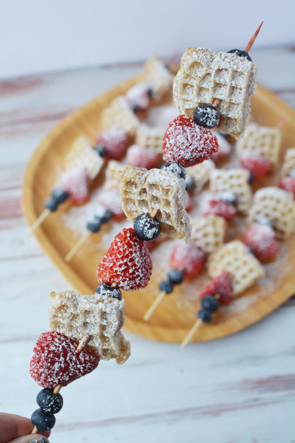 Skewered mini waffles and fruit above a plate full of them.