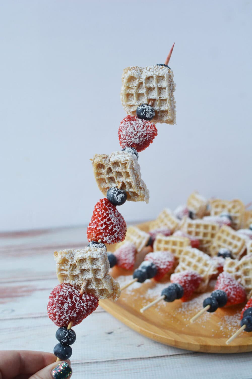 Holding a mini waffle skewer with berries and powdered sugar. 