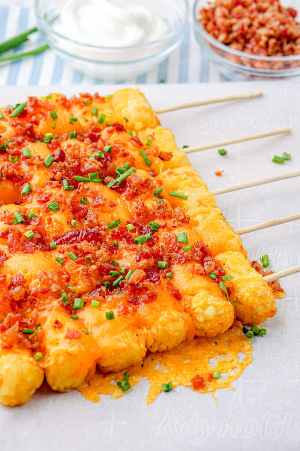 Tater tot skewers next to bowls of sour cream and bacon crumbles